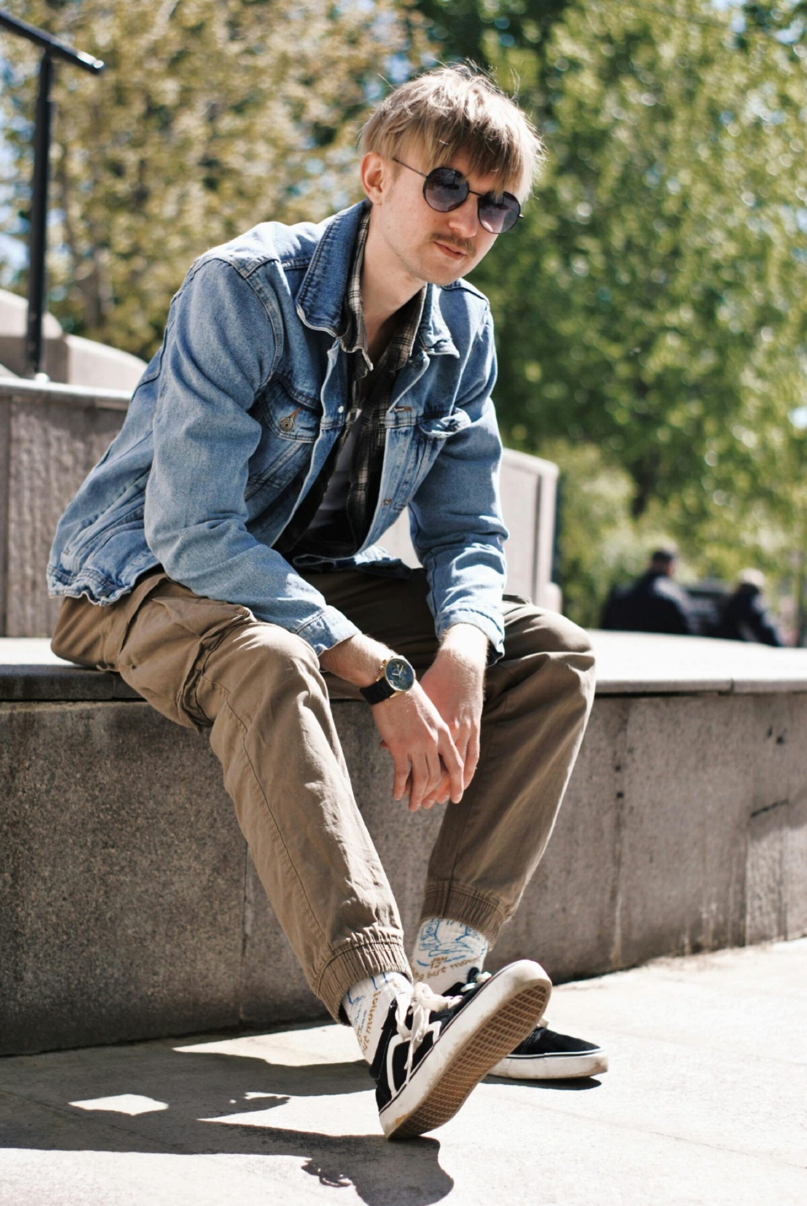 Fashionable young man in sunglasses and a denim jacket relaxing outdoors on a sunny day.