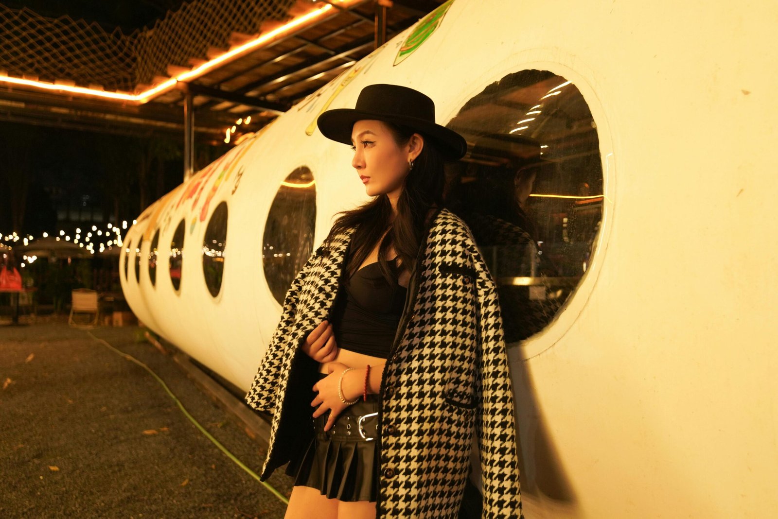 A fashionable woman in a houndstooth coat leans against a tube-shaped structure, illuminated by night lights.